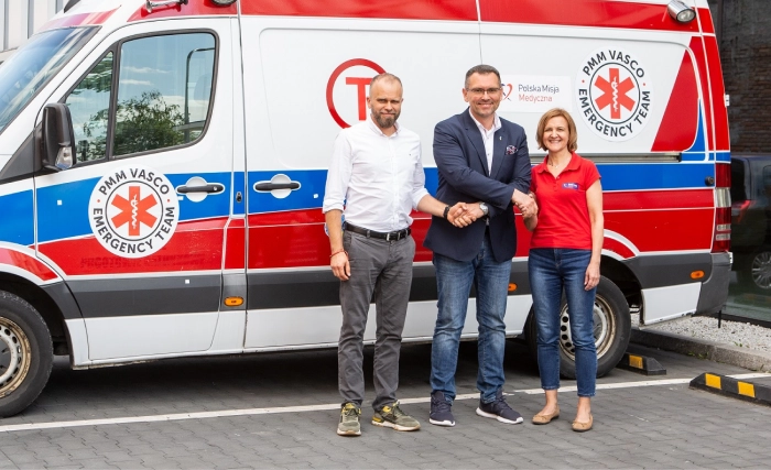 Trzy osoby stoją uśmiechnięte przed biało-czerwono-niebieską karetką zaparkowaną na brukowanej ulicy obok budynku. Dwóch mężczyzn, jeden w garniturze i jeden w białej koszuli, podaje sobie ręce, do których dołącza kobieta w czerwonej koszuli.